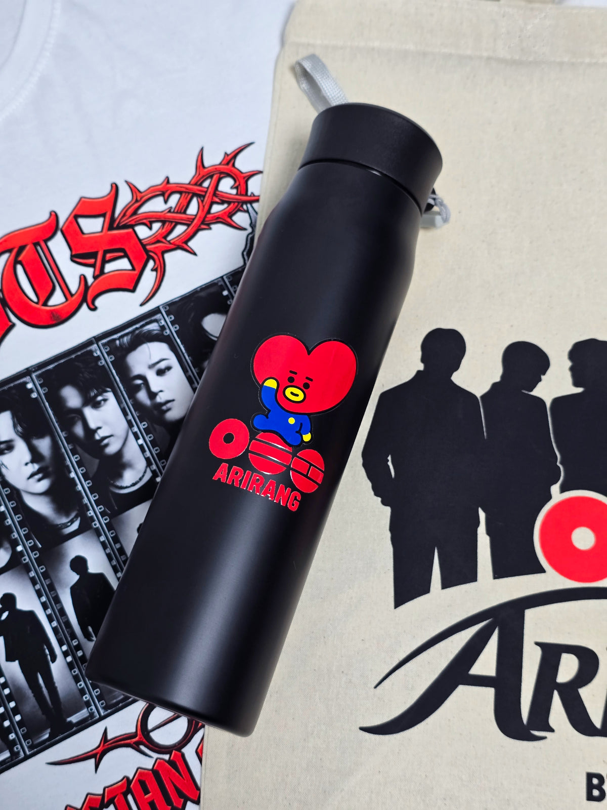 Termo negro de la caja BTS con Tata de BT21 y el logo Arirang, sobre la polera blanca y el totebag con las siluetas de los siete