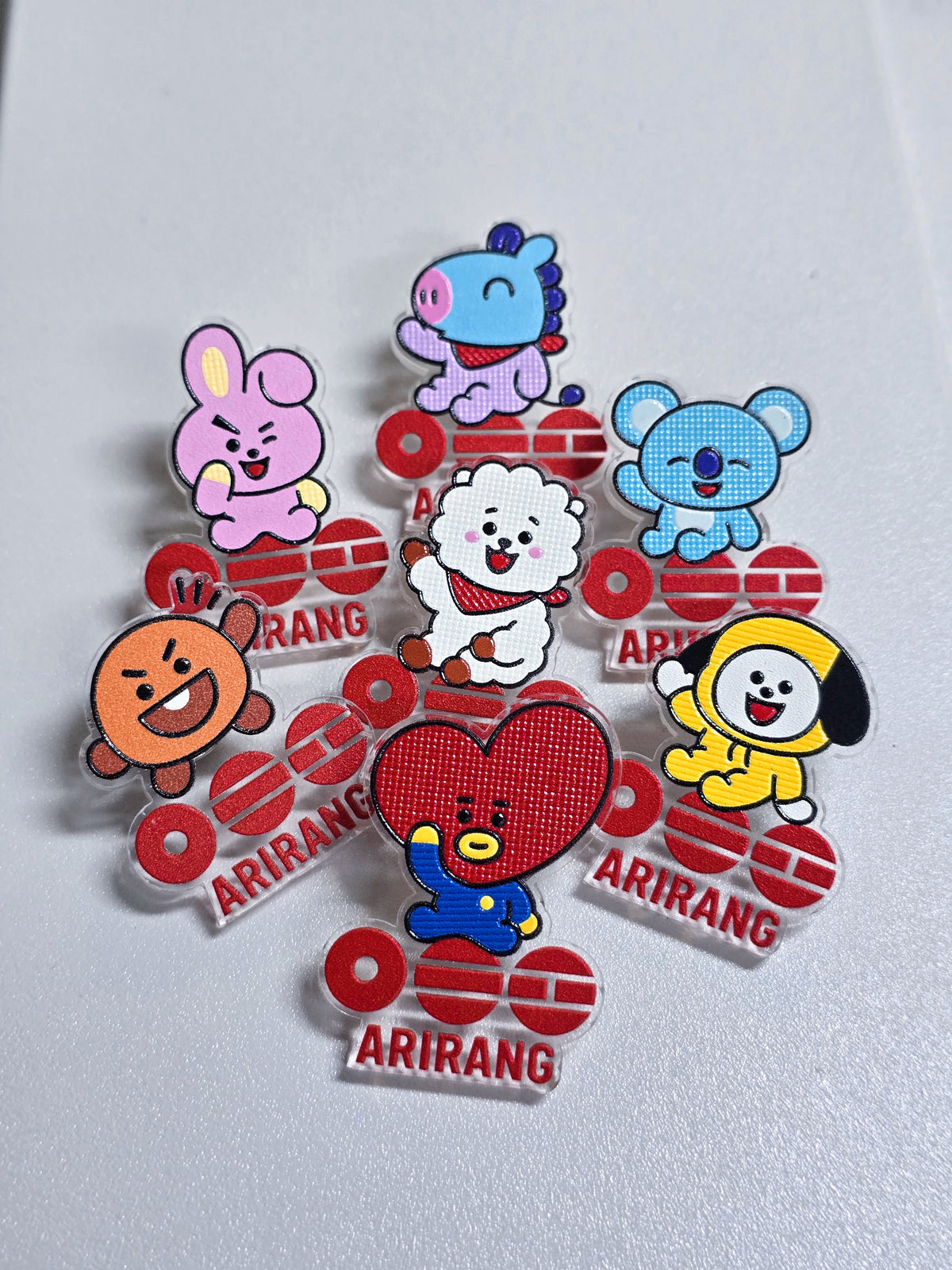 Los siete pines de BT21 de la caja BTS, cada uno con el logo Arirang: Cooky, Mang, Koya, RJ, Shooky, Tata y Chimmy