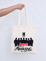 Totebag de tela cruda de la caja BTS, sostenido por una mano: logo BTS, las siluetas de los siete, los círculos Arirang y BTS · 2026
