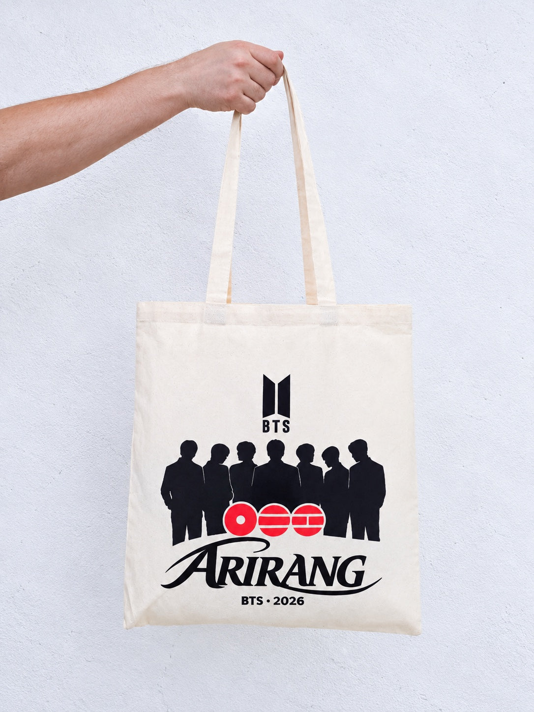 Totebag de tela cruda de la caja BTS, sostenido por una mano: logo BTS, las siluetas de los siete, los círculos Arirang y BTS · 2026