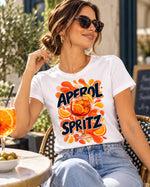 Polera blanca con el diseño Aperol Spritz de la lámina Cócteles, en una terraza con un spritz en la mesa