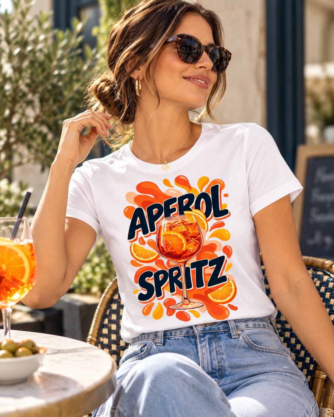 Polera blanca con el diseño Aperol Spritz de la lámina Cócteles, en una terraza con un spritz en la mesa