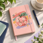 Libreta rosada del Día del Profesor con la manzana floreada sobre pinceladas de colores, en un escritorio con lápiz, café y plantas